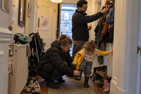 A family getting ready in a hallway: a woman kneels to put a wellington boot on a laughing toddler in a yellow raincoat while a man hangs a child's rucksack on a coat hook.